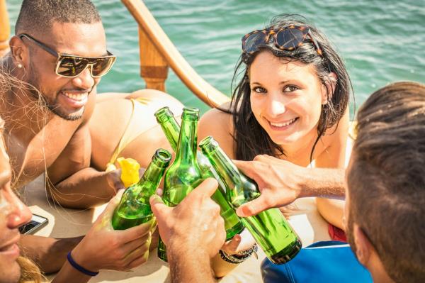 Young adult men and women cl<em></em>inking beer bottles on board boat, woman smiling at camera