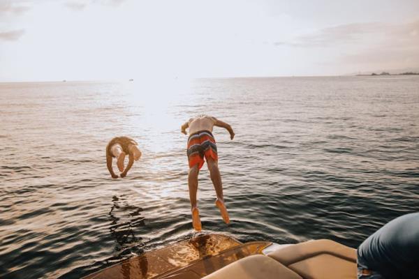 Two young males jumping off boat into calm water, sun setting on horizon